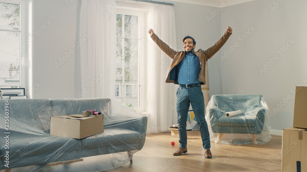 Shot of a Very Happy Man Moves Into His New Apartment and Poses Excited with Raised Arms. Guy Purchased New Home Ready to Start Unpacking Cardboard Boxes.