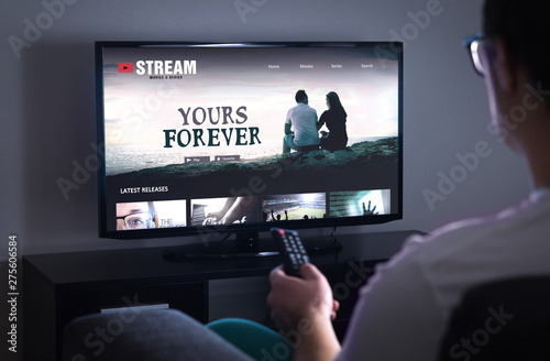 Online movie stream service in smart tv. Streaming series with on demand video (VOD) service in television. Man choosing film to watch with remote. Person sitting on couch at home late at night.