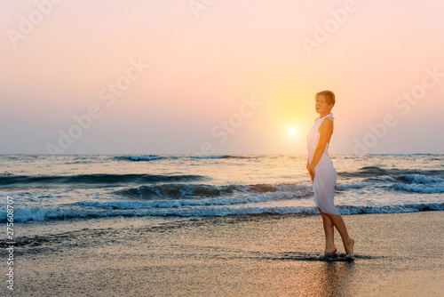 Attractive slender girl in white dress stands in waves on the sea in the light of setting sun. Young woman walks barefoot along the surf and looks into the distance. Rough sea and ripples on water.