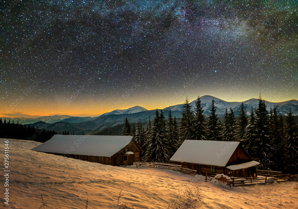 Old wooden house, hut and barn, pile of firewood in deep snow on mountain valley, spruce forest, woody hills on dark starry sky and Milky Way copy space background. Mountain winter night landscape.