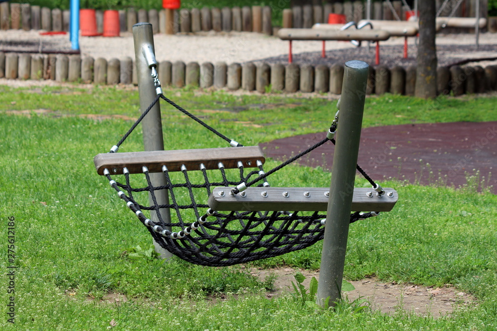 Rope hammock swing enforced with strong wooden and metal support made for climbing and relaxing in local public park surrounded with uncut grass and other playground equipment on warm sunny spring day