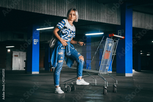 Beautiful girl with short white hair dressed in jeans with shopping carts in the parking lot. Positive young girl in the supermarket trolley.