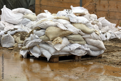 Small pile of white sandbags put on wooden pallet prepared to be put on top of temporary flood protection wall surrounded with mudd and flood water on rainy spring day