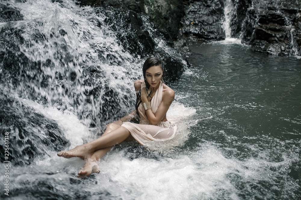 Sensual female in white dress is sitting in the water on a big waterfal