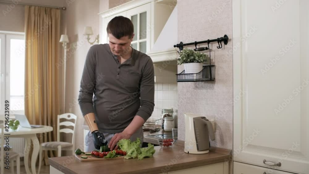 Thigh-up shot of Caucasian man with bionic prosthetic forearm making vegetable salad in kitchen at home, holding knife with artificial hand and cutting cucumber into pieces with precise movements Thigh-up shot of Caucasian man with bionic prosthetic forearm making vegetable salad in kitchen at home, holding knife with artificial hand and cutting cucumber into pieces with precise movements