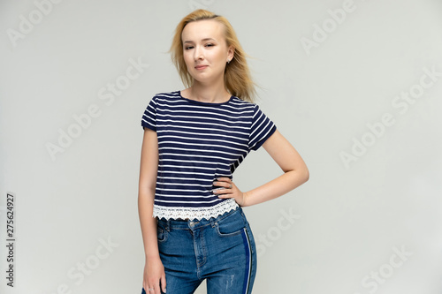 Portrait of a pretty interesting blonde girl on a white background in jeans and a striped blue t-shirt. Standing in front of the camera, smiling, showing hands. Shows a lot of emotions.