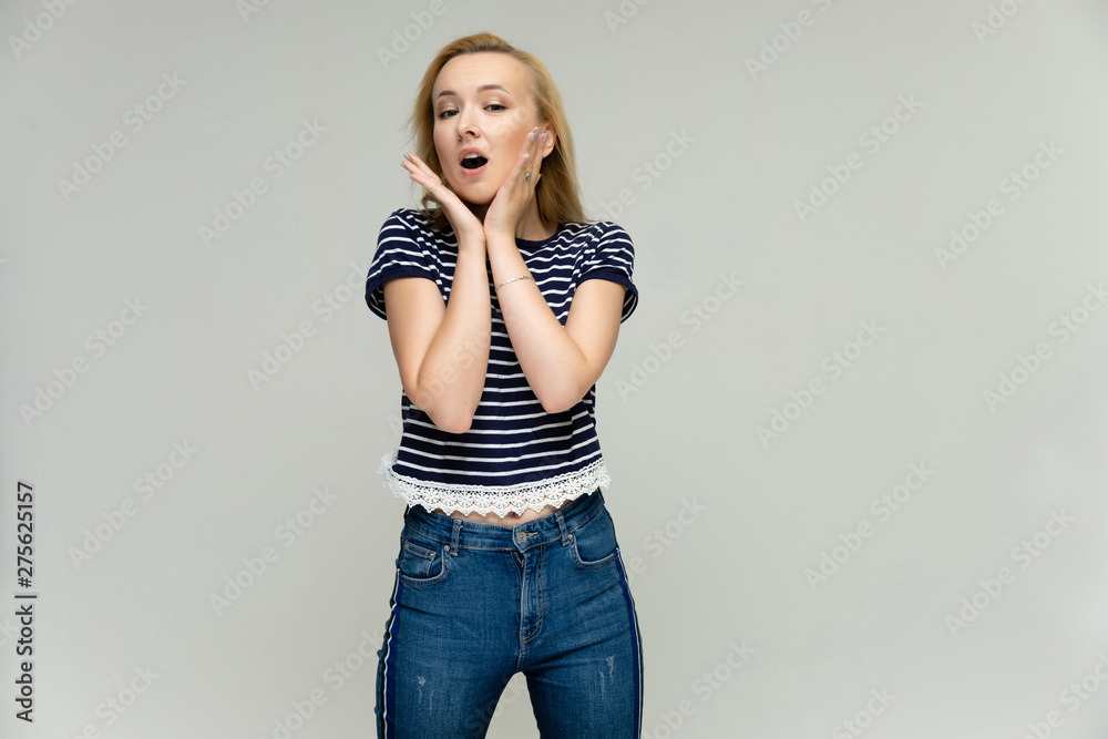 Portrait of a pretty interesting blonde girl on a white background in jeans and a striped blue t-shirt. Standing in front of the camera, smiling, showing hands. Shows a lot of emotions. Portrait of a pretty interesting blonde girl on a white background in jeans and a striped blue t-shirt. Standing in front of the camera, smiling, showing hands. Shows a lot of emotions.