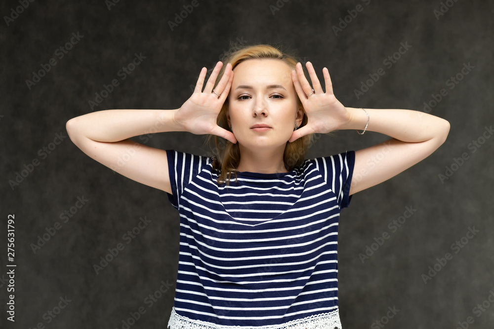 Portrait to the waist of a pretty interesting fashionable blonde girl on a gray background in a striped blue t-shirt. Standing in front of the camera, smiling, showing hands. Shows a lot of emotions. Portrait to the waist of a pretty interesting fashionable blonde girl on a gray background in a striped blue t-shirt. Standing in front of the camera, smiling, showing hands. Shows a lot of emotions.