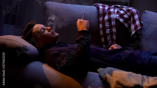 Male teenager smoking cigarette on sofa, risk of fire, awkward age problem