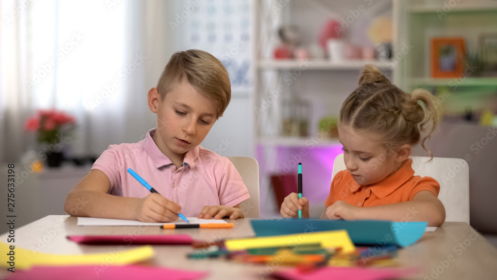 Adorable boy and girl drawing with pencils, sitting at table, kindergarten Adorable boy and girl drawing with pencils, sitting at table, kindergarten