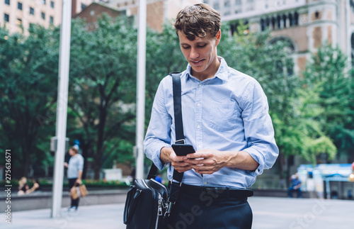 Male entrepreneur walking at urban setting with modern cellphone gadget in hands and typing text message for sending information to partner, businessman chatting via cell in financial district