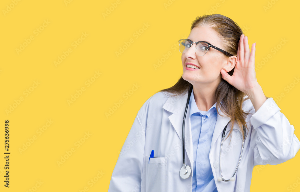 Middle age mature doctor woman wearing medical coat over isolated background smiling with hand over ear listening an hearing to rumor or gossip. Deafness concept.