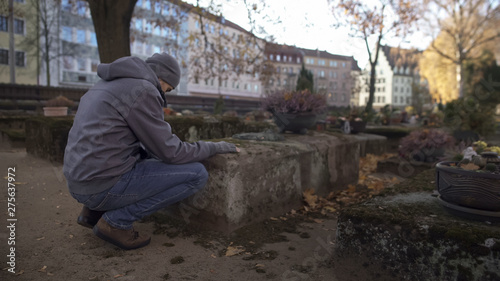 Young man sitting near grave on ancient cemetery praying, mourning for relatives