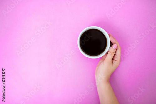 Flatlay with black coffee in a white mug on a pink background. Woman's hand with manicure gently holding cup. Copy space.