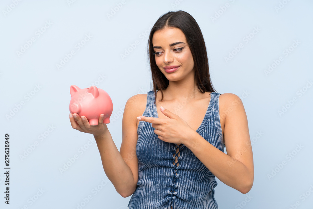 Young woman over isolated blue wall holding a big piggybank Young woman over isolated blue wall holding a big piggybank