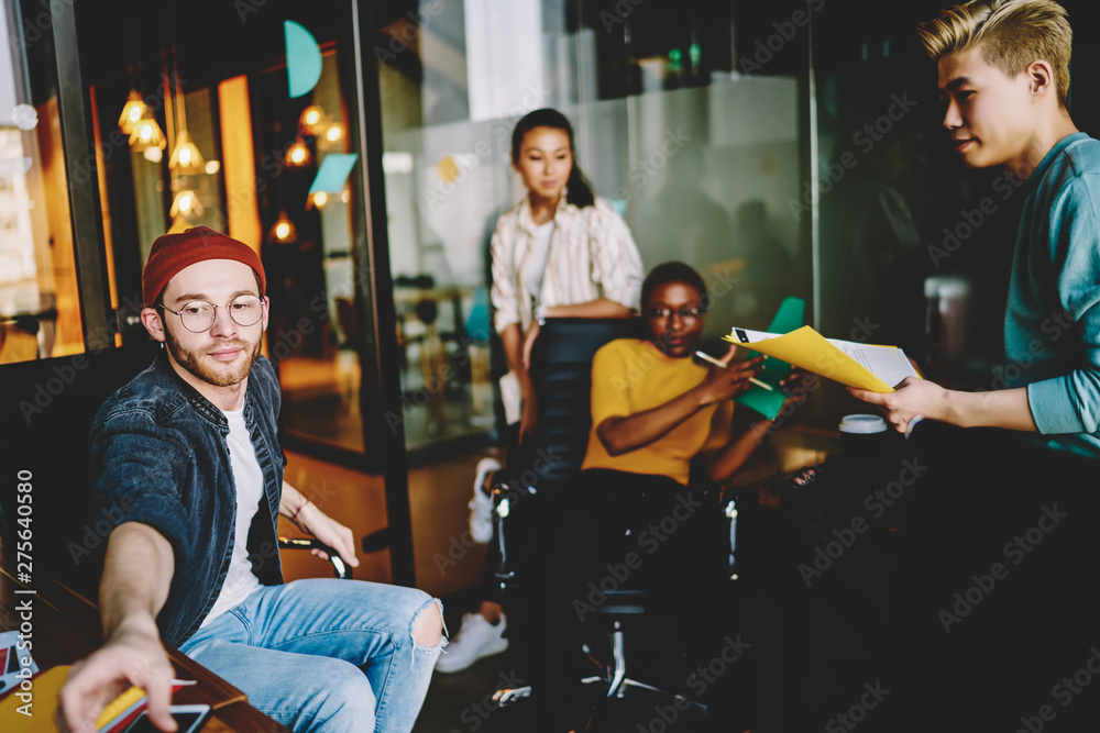 Young hipster guy taking smth from table having meeting with team of employees, skilled multiracial male and female colleagues planning startup strategy together discuss ideas and information.