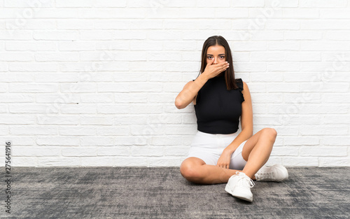 Pretty young woman sitting on the floor covering mouth with hands