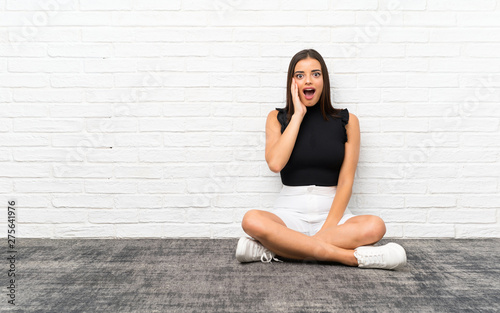 Pretty young woman sitting on the floor with surprise and shocked facial expression
