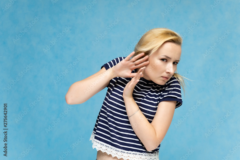 Portrait to the waist of a nice interesting fashionable blonde girl on a blue background in a striped blue t-shirt. Standing in front of the camera, smiling, showing hands. Shows a lot of emotions.