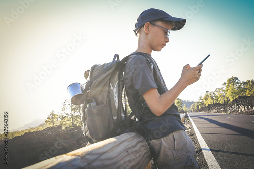 Young boy alone waiting for a ride on the side of the street. Teen with backpack watching new video trends on mobile cell phone and makes social story. Brave adventure and loneliness traveller concept