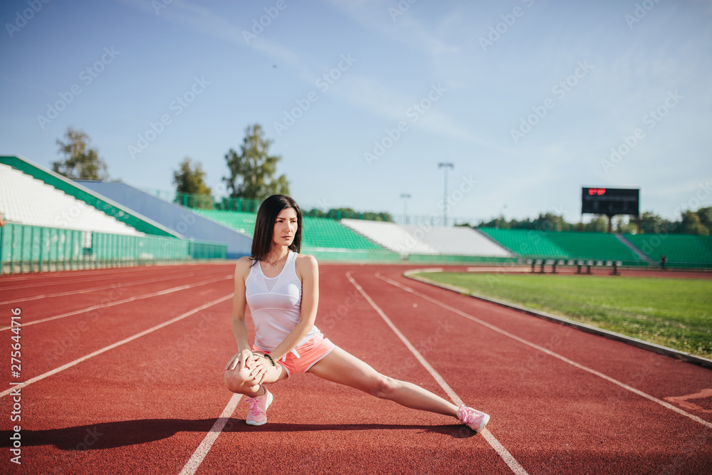 Beautiful fitness brunette girl in sportswear and sneakers does a stretching exercise on the running track at the stadium outdoors. Sun shines onto her body