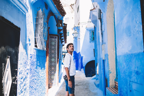 Pensive indian hipster guy standing at antique blue street in old city of Morocco enjoying summer trip for exploring new country, handsome man with retro camera looking around during sightseeing © BullRun