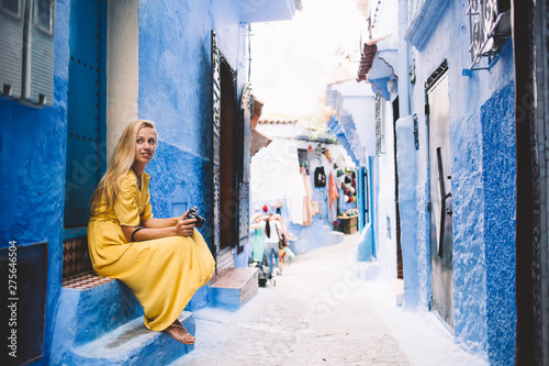 Attractive hipster girl sitting near blue wall in colorful street - Morocco thinking about unesco heritage, young female in yellow dress with camera taking rest in berber medina on vacation © BullRun