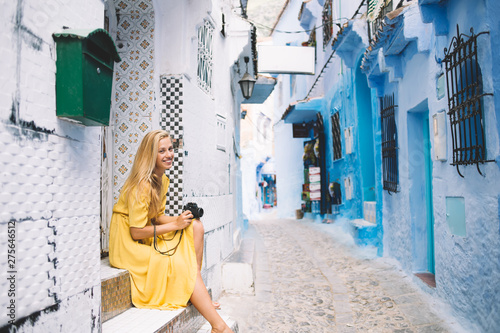 Charming woman sitting near oriental mosaic on ethnic wall - Morocco smiling at camera on summer journey, portrait of blond girl with retro equipment enjoying about architecture in old berber heritage © BullRun