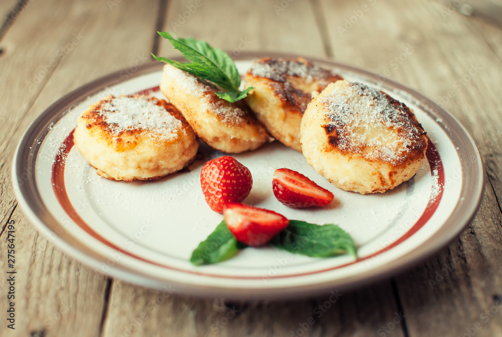 Gourmet breakfast - cottage cheese pancakes, cheesecakes, cottage cheese pancakes with strawberries, mint and powdered sugar in a white plate. Useful dessert on a wooden table in rustic style. Selecti