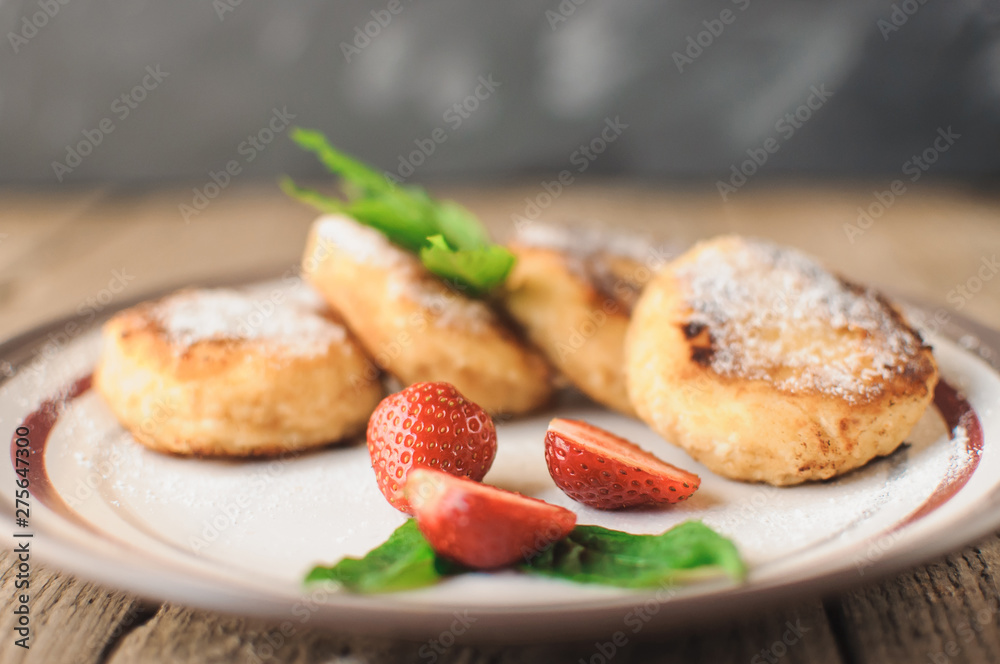 Gourmet breakfast - cottage cheese pancakes, cheesecakes, cottage cheese pancakes with strawberries, mint and powdered sugar in a white plate. Useful dessert on a wooden table in rustic style. Selecti