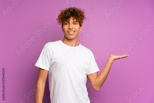 Young african american man over isolated purple wall holding copyspace imaginary on the palm to insert an ad