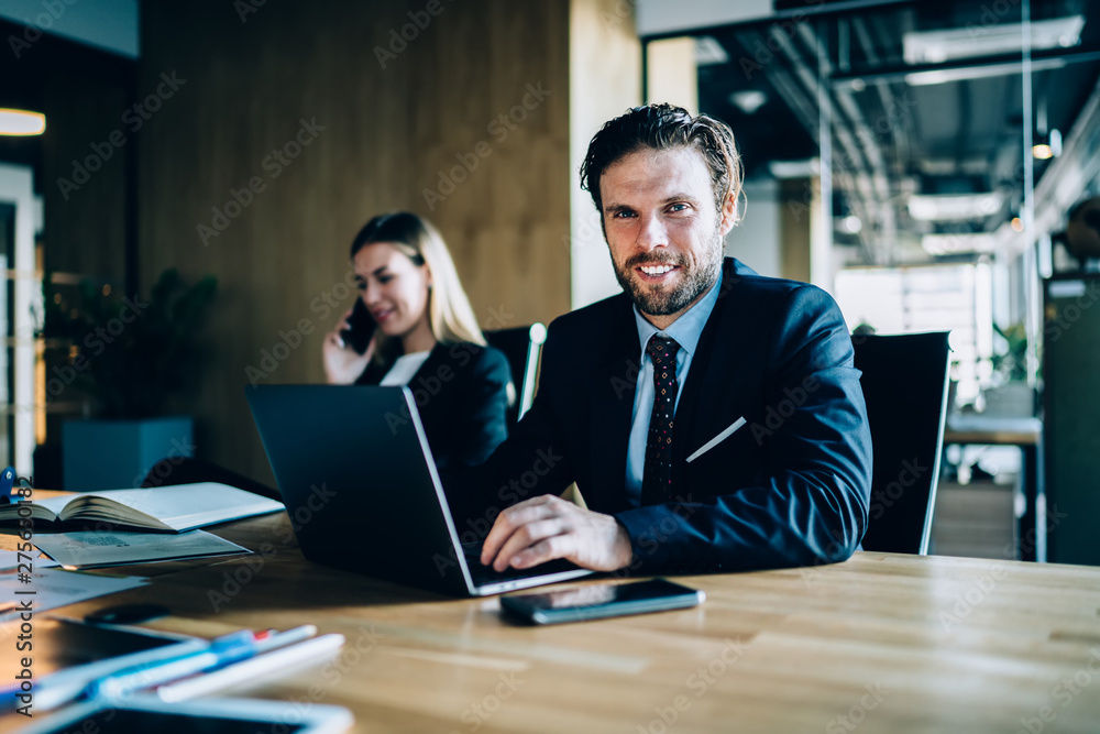 Portrait of successful cheerful male office worker dressed in formal suit smiling at camera while sitting at desktop with modern technology for browsing internet on financial websites connected to 4g