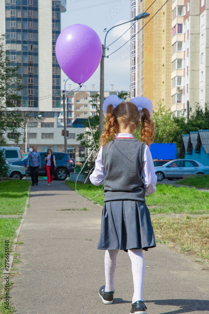A girl with large white bows on her head, a beautiful hairdo in a school uniform, is walking along the sidewalk from the school. She is wearing a school uniform. View from the back