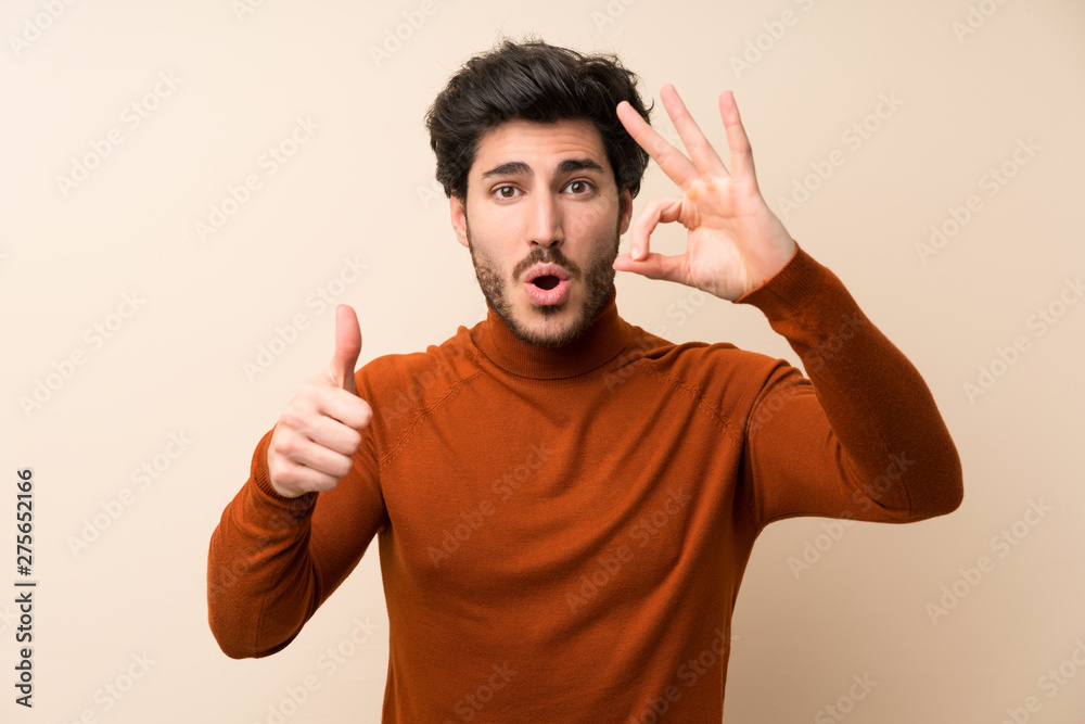 Handsome over isolated wall showing ok sign and thumb up gesture Handsome over isolated wall showing ok sign and thumb up gesture