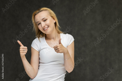 Waist up portrait of pretty interesting fashionable blonde girl on gray background in white t-shirt. Standing in front of the camera, smiling, showing hands. Shows a lot of emotions.