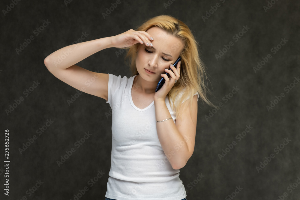 Fototapeta premium Waist up portrait of pretty interesting fashionable blonde girl on gray background in white t-shirt with smartphone. Standing in front of the camera, smiling, showing hands. many emotions.