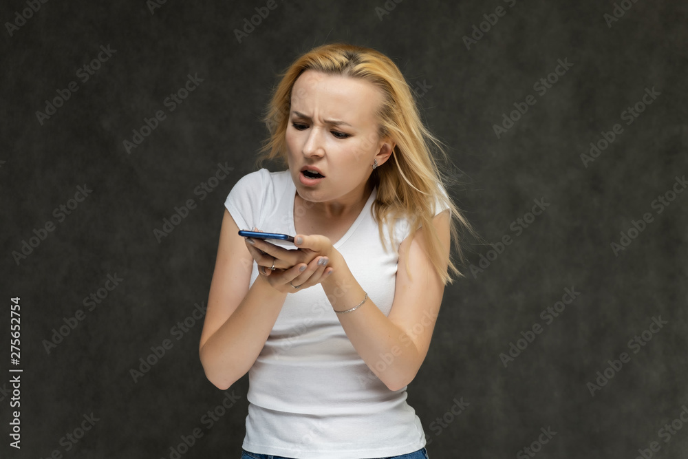 Obraz premium Waist up portrait of pretty interesting fashionable blonde girl on gray background in white t-shirt with smartphone. Standing in front of the camera, smiling, showing hands. many emotions.