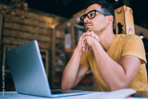 Contemplative young male student in spectacles for vision correction looking away and pondering thinking on idea for university project, pensive hipster guy sitting at desktop with modern laptop