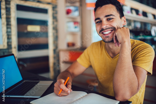 Portrait of happy hipster guy looking at camera and smiling while sitting at desktop with laptop computer for e learning and textbook for knowledge education, cheerful male student feeling good