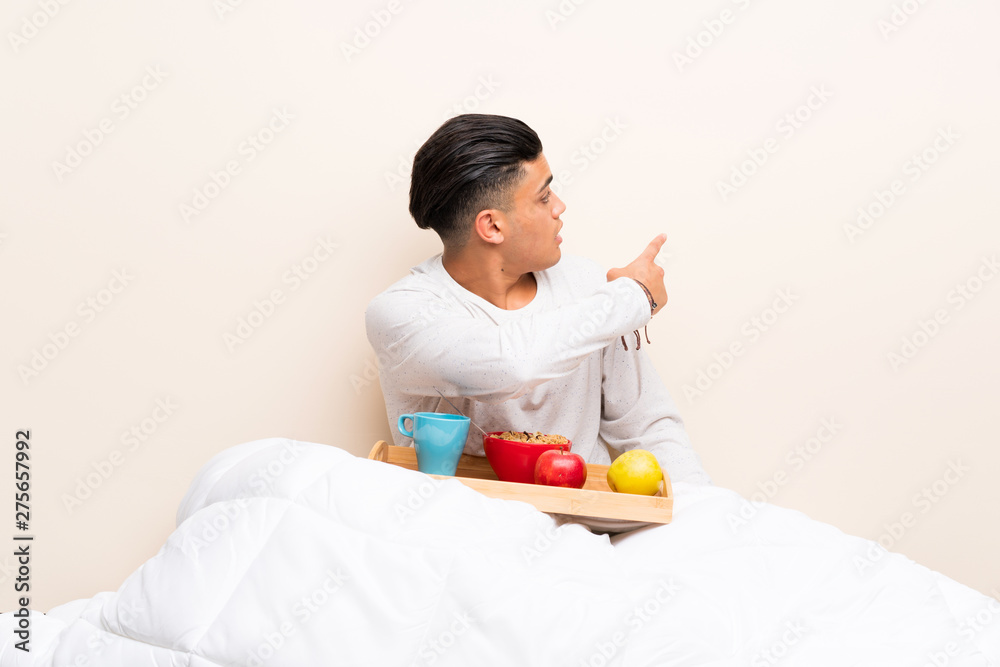 Young man having breakfast in bed pointing back with the index finger Young man having breakfast in bed pointing back with the index finger