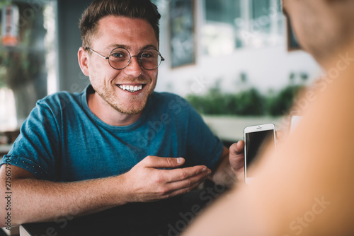 Portrait of cheerful hipster guy in optical spectacles holding modern cell device and smiling at camera while spending leisure time with best friend for discussing notifications, millennial people