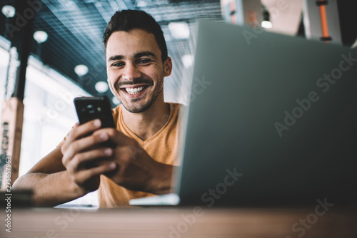 Portrait of happy hipster blogger with modern mobile phone in hands smiling at camera while laughing from funny joke on website, millennial cheerful man with digital technologies enjoying leisure time photo