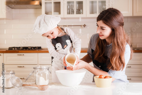 The little daughter in the chef's hat and apron and her mother prepare baking in the bright, classic kitchen.