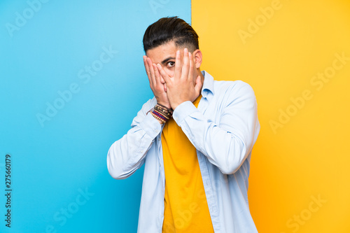 Young man over isolated colorful background covering eyes and looking through fingers
