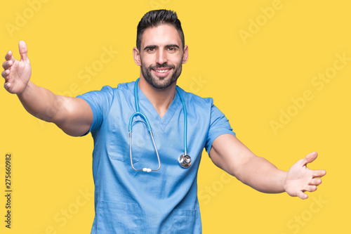 Handsome young doctor surgeon man over isolated background looking at the camera smiling with open arms for hug. Cheerful expression embracing happiness.