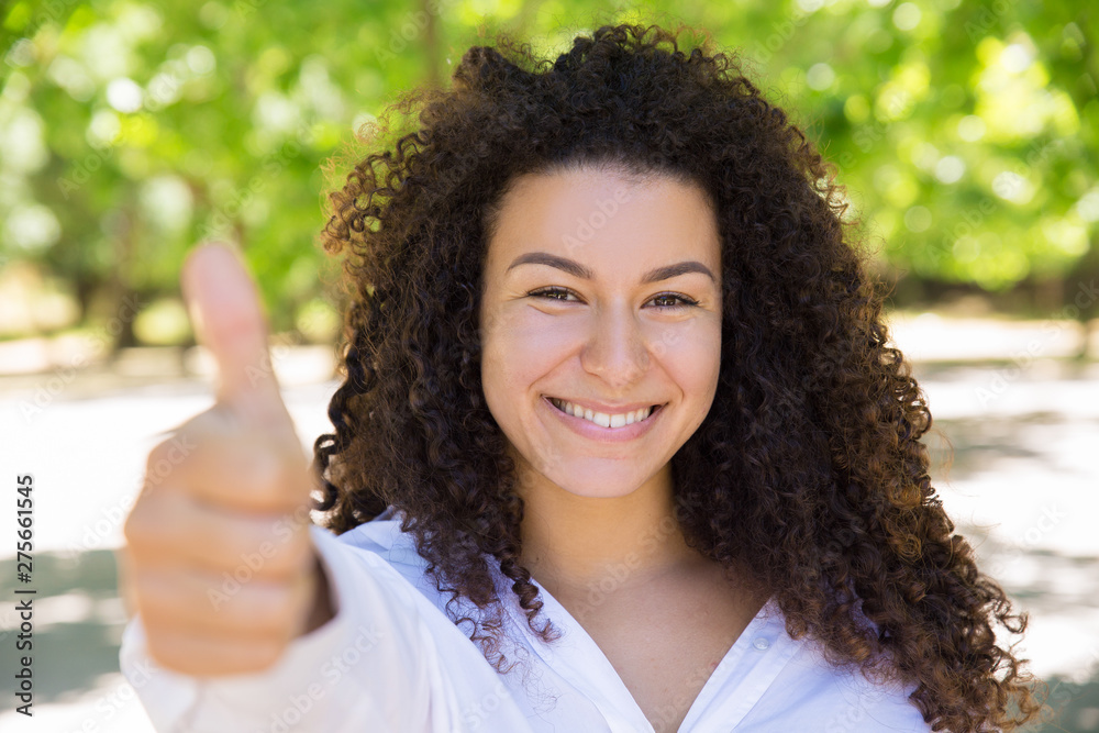 Smiling pretty curly-haired lady showing thumb up in park. Beautiful young woman wearing blouse and looking at camera with blurred green trees in background. Promotion concept. Front view. Smiling pretty curly-haired lady showing thumb up in park. Beautiful young woman wearing blouse and looking at camera with blurred green trees in background. Promotion concept. Front view.