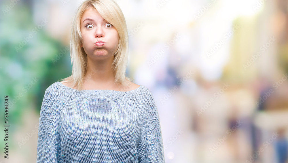 Young beautiful blonde woman wearing winter sweater over isolated background puffing cheeks with funny face. Mouth inflated with air, crazy expression.