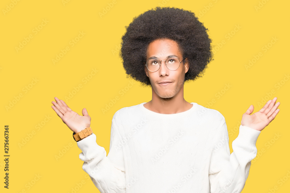 Young african american man with afro hair wearing glasses clueless and confused expression with arms and hands raised. Doubt concept. Young african american man with afro hair wearing glasses clueless and confused expression with arms and hands raised. Doubt concept.