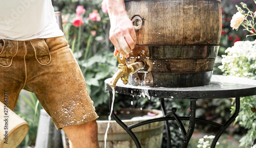 bayerischer Mann in Lederhose sticht ein Holzfass Bier im Garten an und genießt den ersten Schluck photo