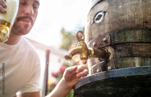 bayerischer Mann in Lederhose sticht ein Holzfass Bier im Garten an und genießt den ersten Schluck photo